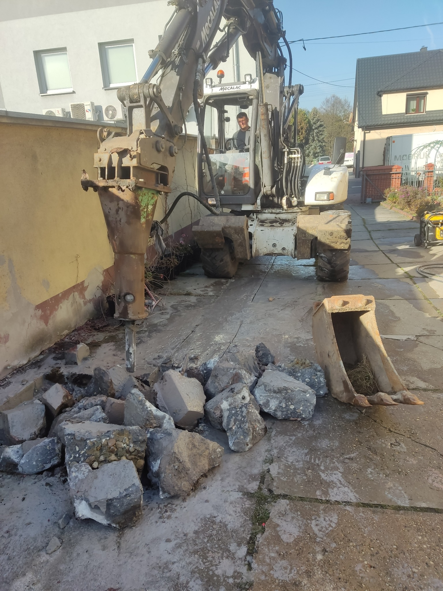 Operator w kabinie koparki z hydraulicznym młotem pneumatycznym kruszy beton na terenie posesji w Mysłowicach. Obok leży sterta gruzu i łyżka koparki.