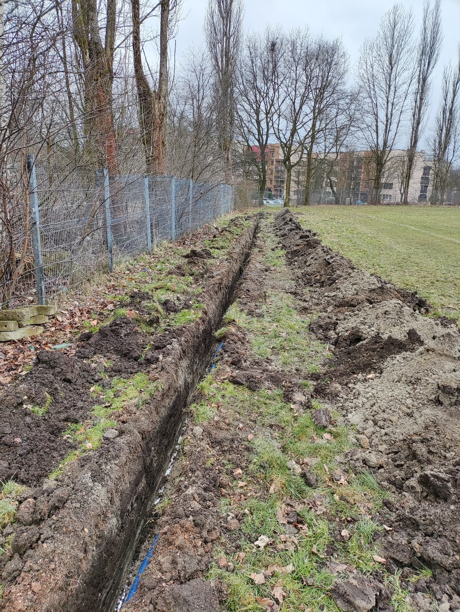 Długi, wąski wykop w ziemi na trawiastym terenie, zabezpieczony niebieską rurą karbowaną, widoczny fragment ogrodzenia z siatki i drzewa bezlistne w tle.