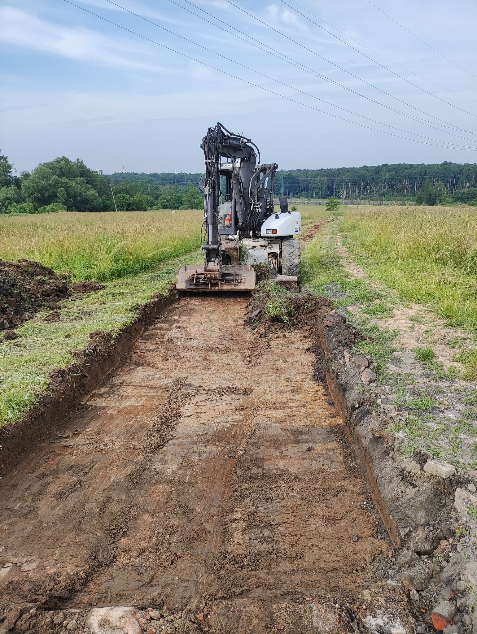 Koparka kołowa w trakcie wykonywania wykopu liniowego w terenie trawiastym, widoczne ślady pracy maszyny na dnie wykopu, w tle drzewa i słupy energetyczne.