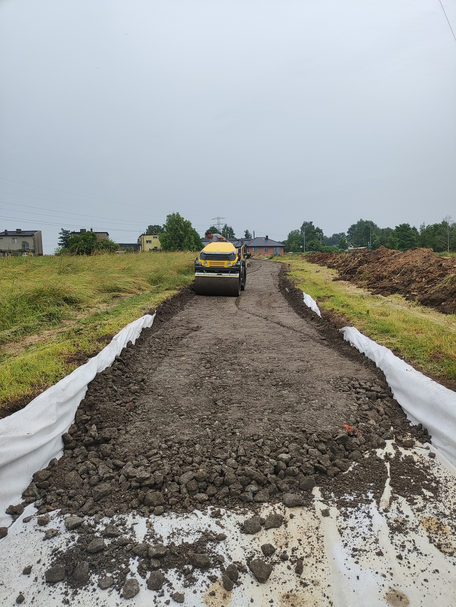 Żółty walec drogowy ubija nawierzchnię drogi gruntowej, wyłożonej białą geowłókniną, w tle zabudowania wiejskie i pochmurne niebo.