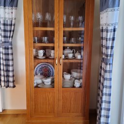 Zakład Stolarski ASKO Joanna Kołodziejska - Drewniana witryna z przeszklonymi drzwiami, wypełniona porcelaną i szkłem, stojąca na parkiecie, z zasłonami w kratę po bokach.