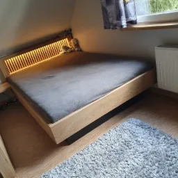 Drewniane łóżko z podświetlanym zagłówkiem z lameli, ustawione na poddaszu, z pluszowymi zabawkami i lampką nocną na szafce obok, oraz puszystym dywanem na podłodze.