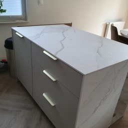 Zakład Stolarski ASKO Joanna Kołodziejska - Wyspa kuchenna z szarymi frontami i blatem imitującym marmur, zlokalizowana w Rożno-Parcele, z widocznymi trzema szufladami z podłużnymi, jasnymi uchwytami.