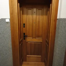 Drewniane drzwi wejściowe do mieszkania z numerem 4, osadzone w drewnianej futrynie, widoczne z bliska na tle szarej ściany klatki schodowej.