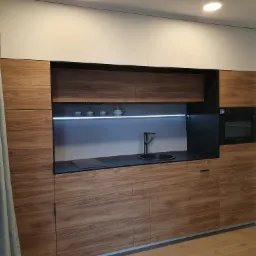 Nowoczesna kuchnia z drewnianymi frontami, białymi górnymi szafkami, czarnym blatem i szklanymi luksferami w tle.