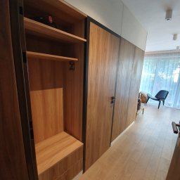 Zakład Stolarski ASKO Joanna Kołodziejska - Wnętrze przedpokoju z zabudowaną szafą przesuwną z drewnianymi frontami i czarnymi obramowaniami, częściowo otwarta z widocznymi półkami i szufladami, na górnej półce leży czarny akumulatorowy...