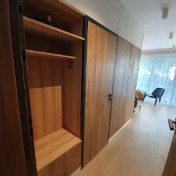 Wnętrze przedpokoju z zabudowaną szafą przesuwną z drewnianymi frontami i czarnymi obramowaniami, częściowo otwarta z widocznymi półkami i szufladami, na górnej półce leży czarny akumulatorowy...