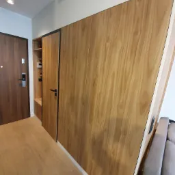 Drewniana zabudowa w przedpokoju z szafkami i półkami, fronty z wyraźnym usłojeniem, drzwi wejściowe po lewej stronie, podłoga z paneli w odcieniach beżu.