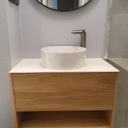 Nowoczesna łazienka z okrągłą białą umywalką nablatową, armaturą w kolorze grafitowym i szafką z jasnego drewna z otwartą półką, odbicie w okrągłym lustrze.