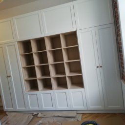 Zakład Stolarski ASKO Joanna Kołodziejska - Biała zabudowa meblowa z otwartymi półkami, widoczne zabezpieczenia podłogi podczas montażu, fragment ściany z cegły przy oknie.