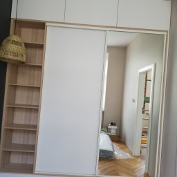 Zakład Stolarski ASKO Joanna Kołodziejska - Biała szafa wnękowa z przesuwnymi drzwiami lustrzanymi, zintegrowana z regałem na książki z jasnego drewna, uzupełniona o wiszącą lampę z naturalnego materiału.