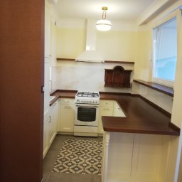 Zakład Stolarski ASKO Joanna Kołodziejska - Jasna kuchnia z drewnianymi blatami i wzorzystą podłogą, widok na białą kuchenkę gazową i okno z drewnianym parapetem.