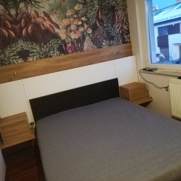 Zakład Stolarski ASKO Joanna Kołodziejska - Sypialnia z drewnianymi szafkami nocnymi i zagłówkiem z motywem botanicznym na tapecie, widok z okna na ośnieżony dach sąsiedniego domu.