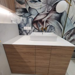 Zakład Stolarski ASKO Joanna Kołodziejska - Nowoczesna łazienka z drewnianą szafką pod umywalką, białym blatem i umywalką nablatową. Na ścianie tapeta w kwiaty, złota armatura i designerskie oświetlenie.