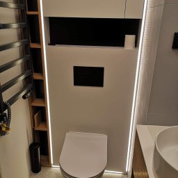 Zakład Stolarski ASKO Joanna Kołodziejska - Nowoczesna łazienka z podświetleniem LED: wisząca toaleta, grzejnik drabinkowy z drewnianymi półkami, umywalka nablatowa i zabudowa meblowa w bieli i drewnie.