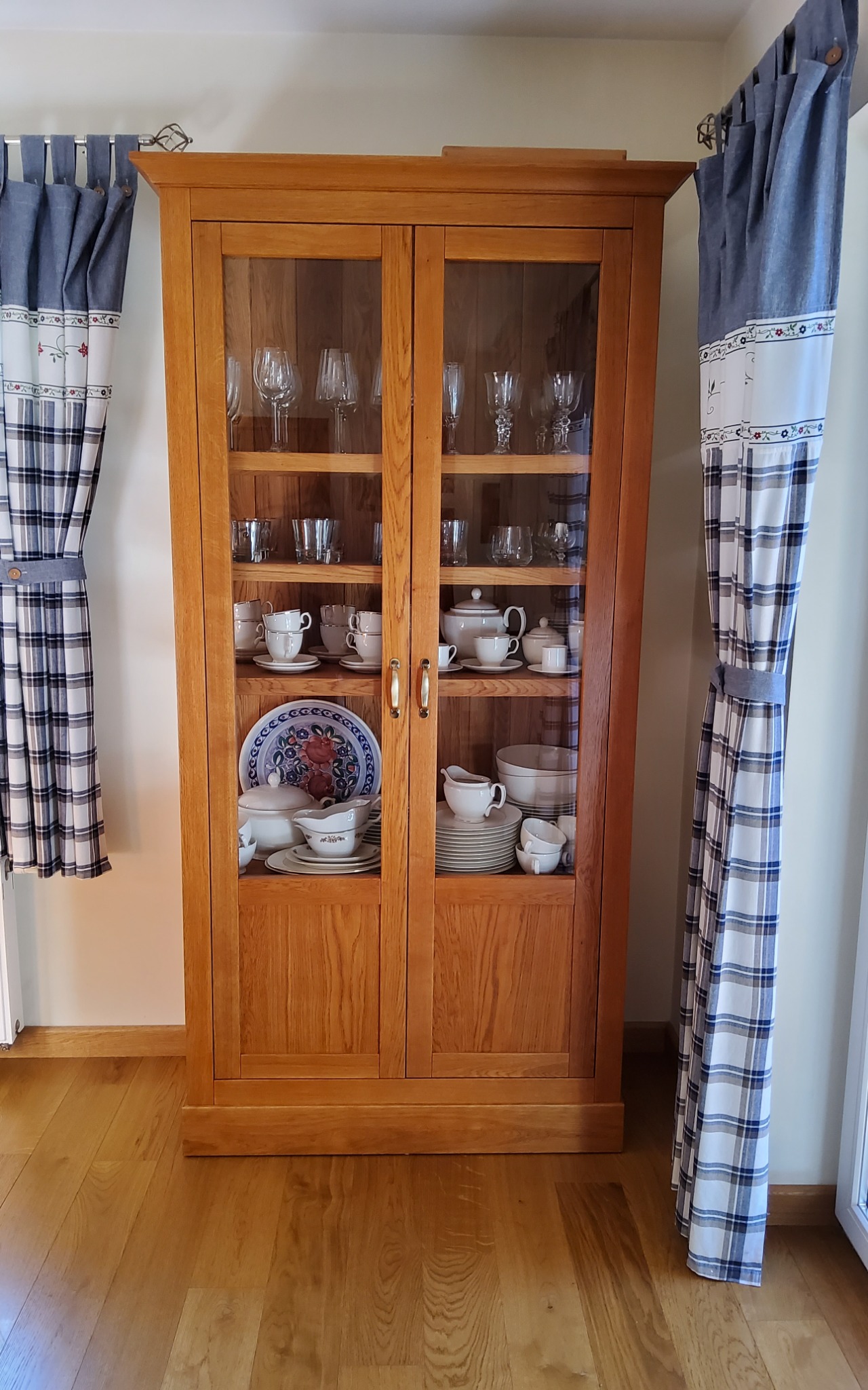 Drewniana witryna z przeszklonymi drzwiami, wypełniona porcelaną i szkłem, stojąca na parkiecie, z zasłonami w kratę po bokach.