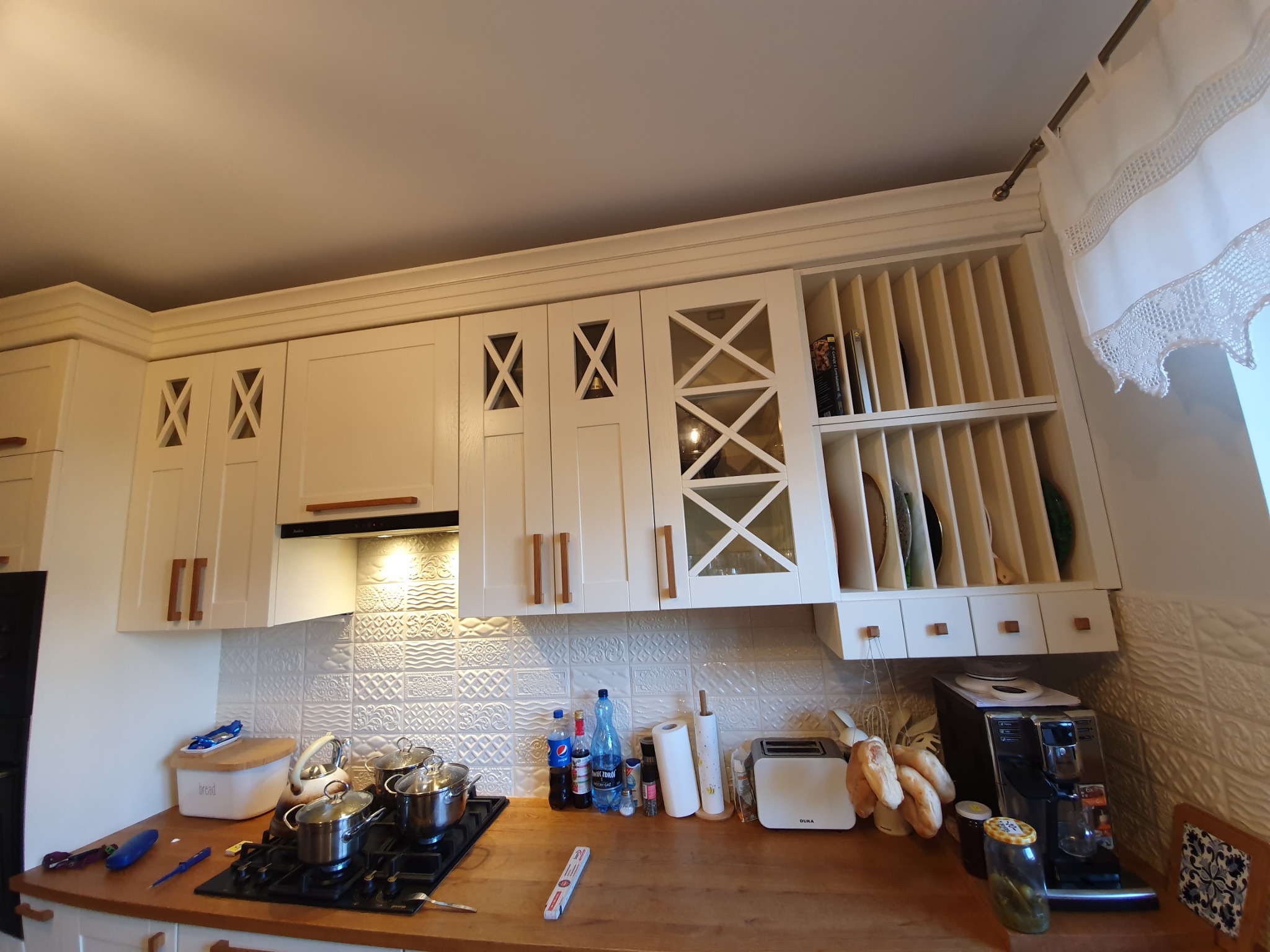 Jasna zabudowa kuchenna z frezowanymi frontami i przeszklonymi szafkami, widok na blat z elementami wyposażenia i akcesoriami kuchennymi.