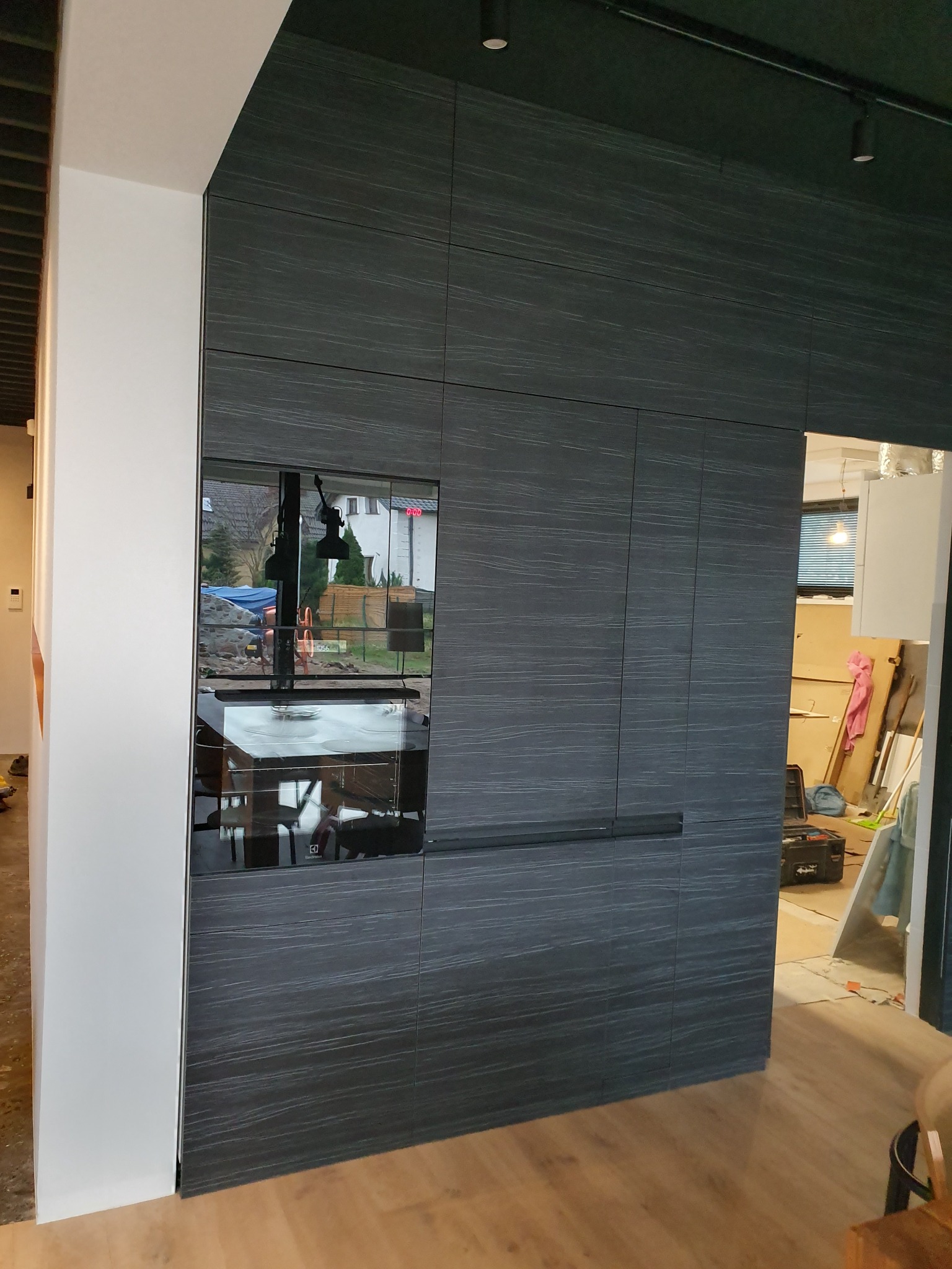 Ciemna zabudowa kuchenna z fornirem o wyraźnym usłojeniu, wkomponowana piekarnik, widoczne elementy instalacji w sąsiednim pomieszczeniu.