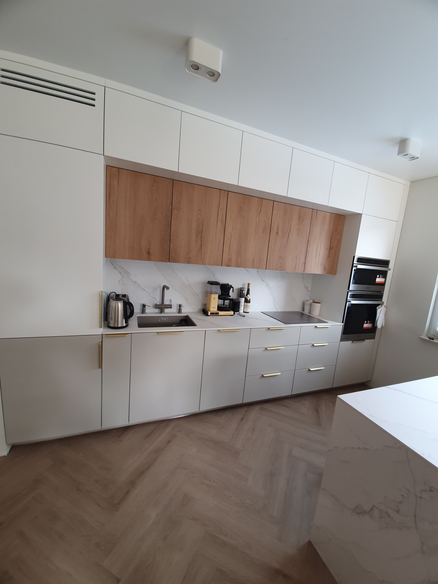 Nowoczesna kuchnia z białymi i szarymi szafkami, drewnianymi akcentami nad blatem, marmurowym backsplashiem i podłogą w jodełkę.