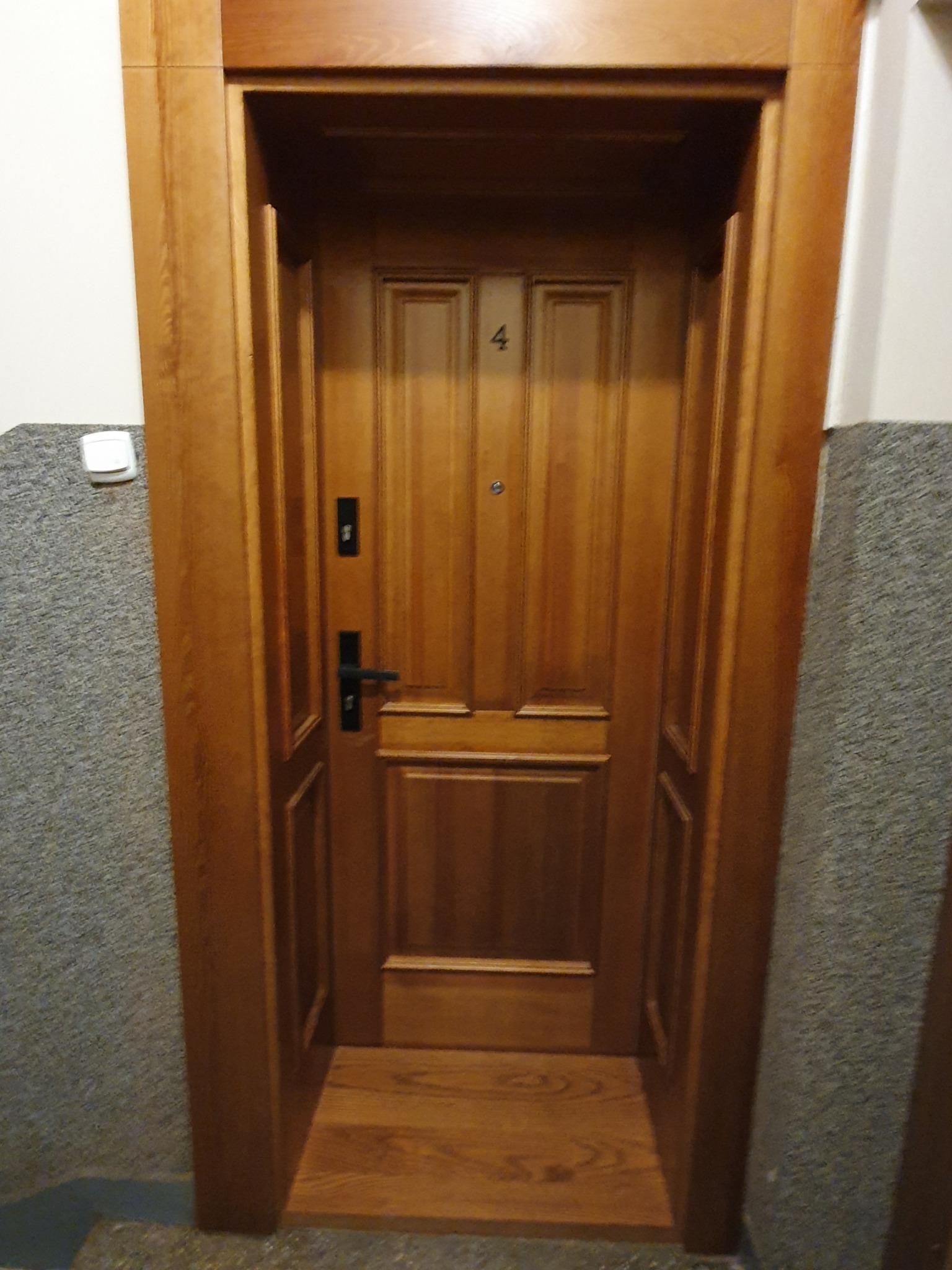 Drewniane drzwi wejściowe do mieszkania z numerem 4, osadzone w drewnianej futrynie, widoczne z bliska na tle szarej ściany klatki schodowej.