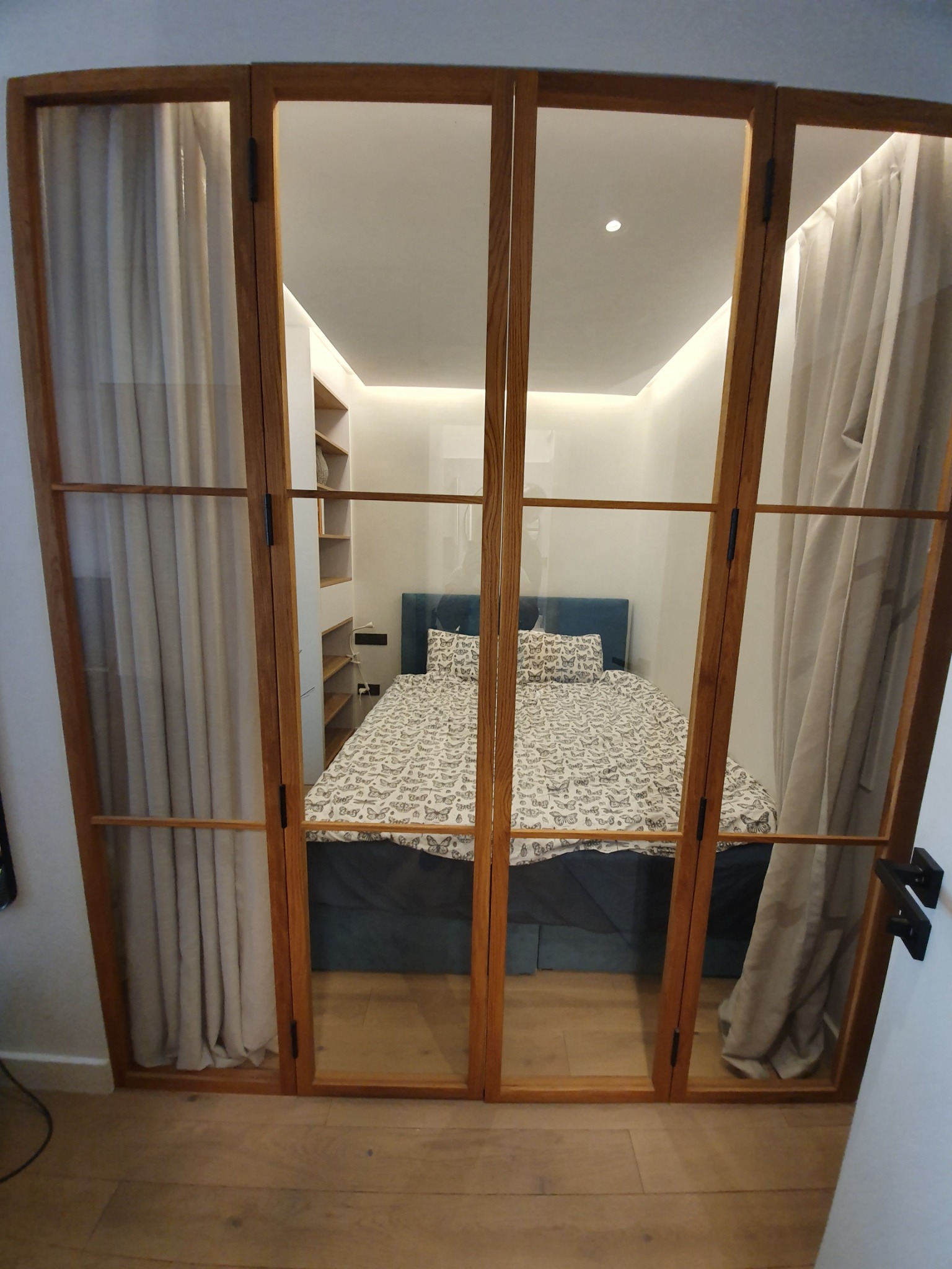 Drewniane drzwi wewnętrzne z szybami, oddzielające sypialnię z łóżkiem w niebieskiej ramie i pościelą w motyle, oraz zasłony w kolorze beżowym.