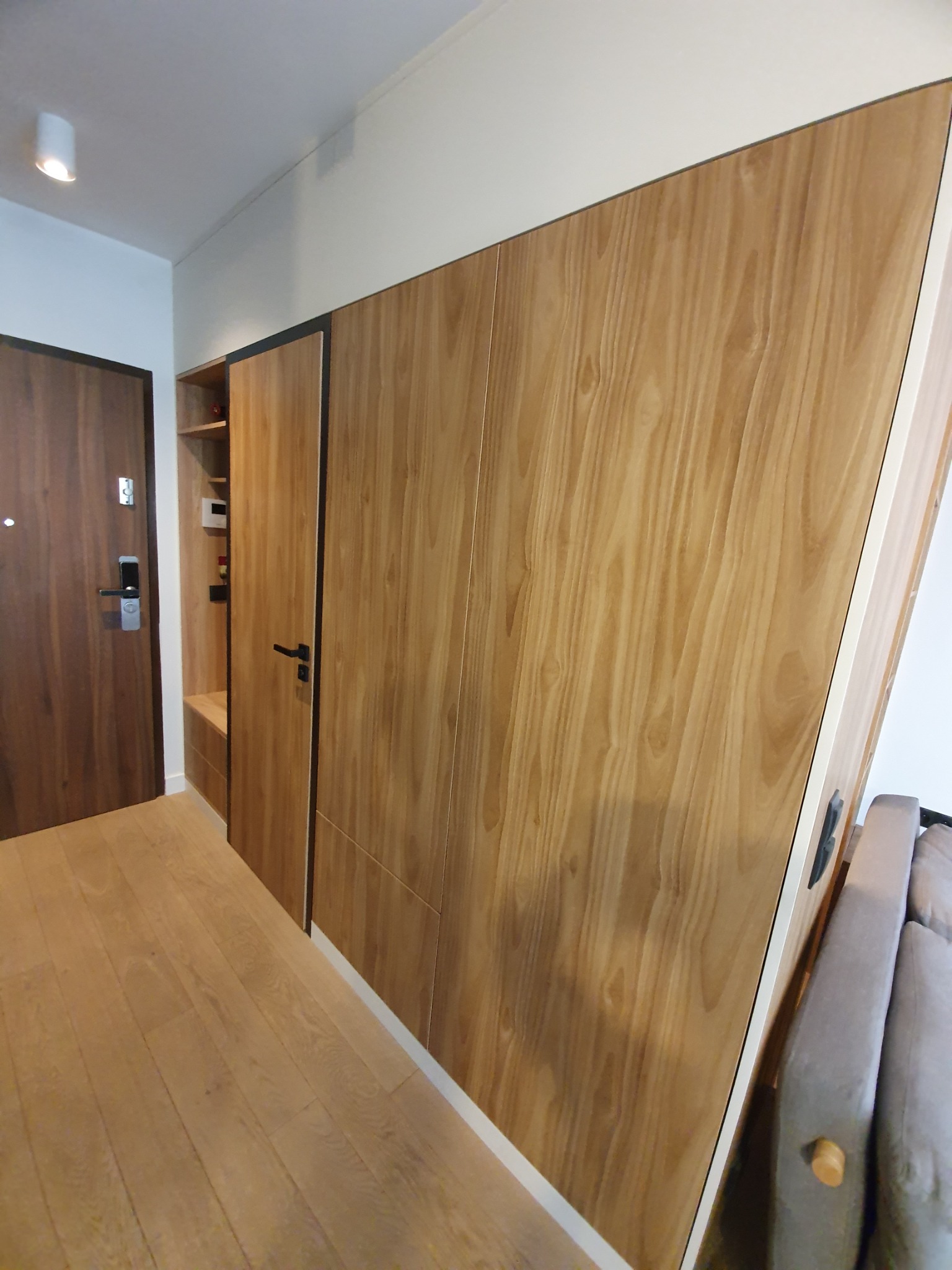 Drewniana zabudowa w przedpokoju z szafkami i półkami, fronty z wyraźnym usłojeniem, drzwi wejściowe po lewej stronie, podłoga z paneli w odcieniach beżu.