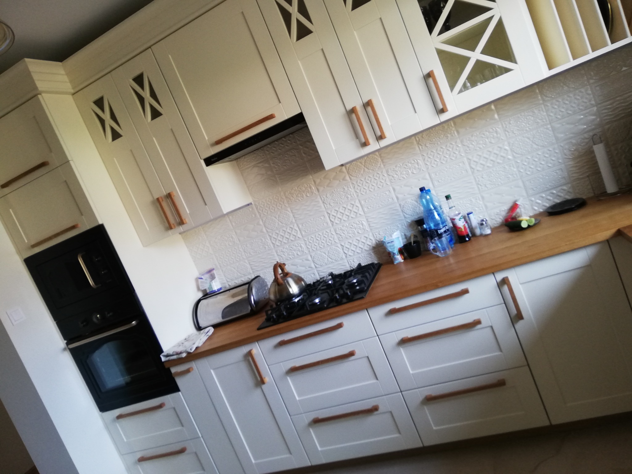 Jasna kuchnia z białymi szafkami z drewnianymi uchwytami, drewniany blat, płyta gazowa, czajnik i piekarnik w zabudowie.