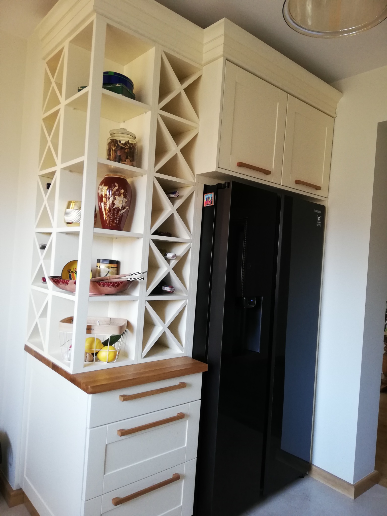 Biała zabudowa kuchenna z regałem na wino i szafkami nad czarną lodówką, widoczne drewniane uchwyty szuflad i szafek, na półkach dekoracyjne naczynia i owoce.