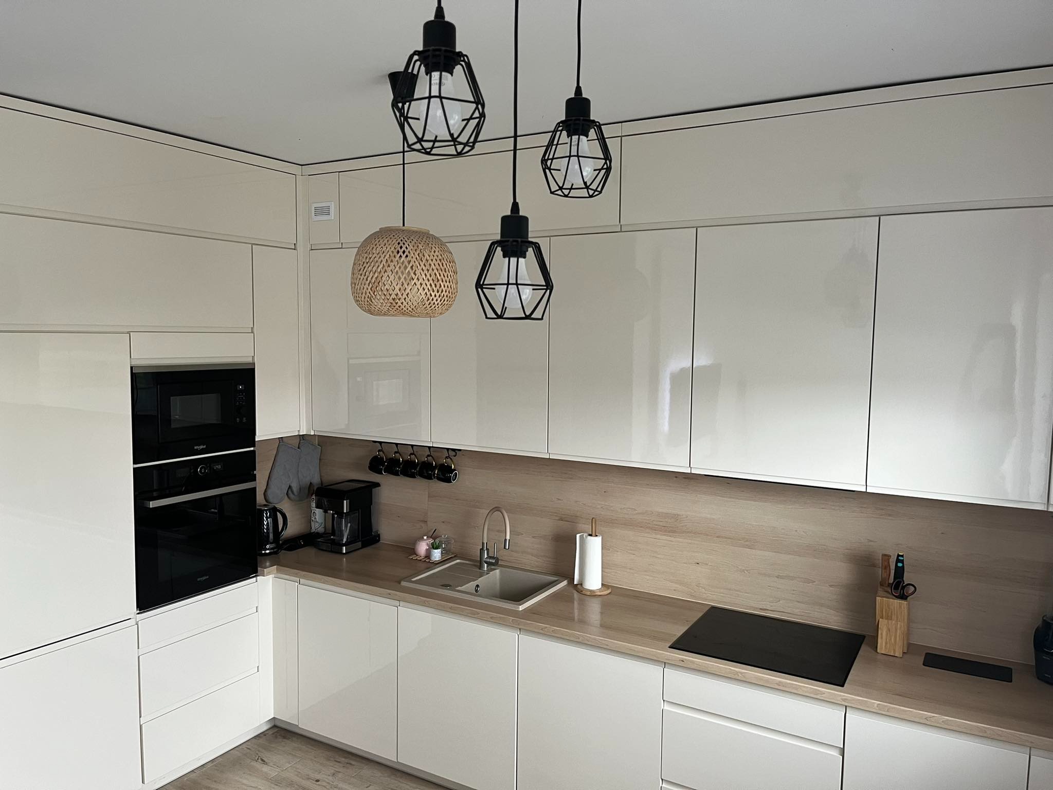 Nowoczesna, jasna kuchnia z błyszczącymi frontami, drewnianym blatem i trzema wiszącymi lampami o geometrycznych kloszach, z których jedna jest pleciona z wikliny.
