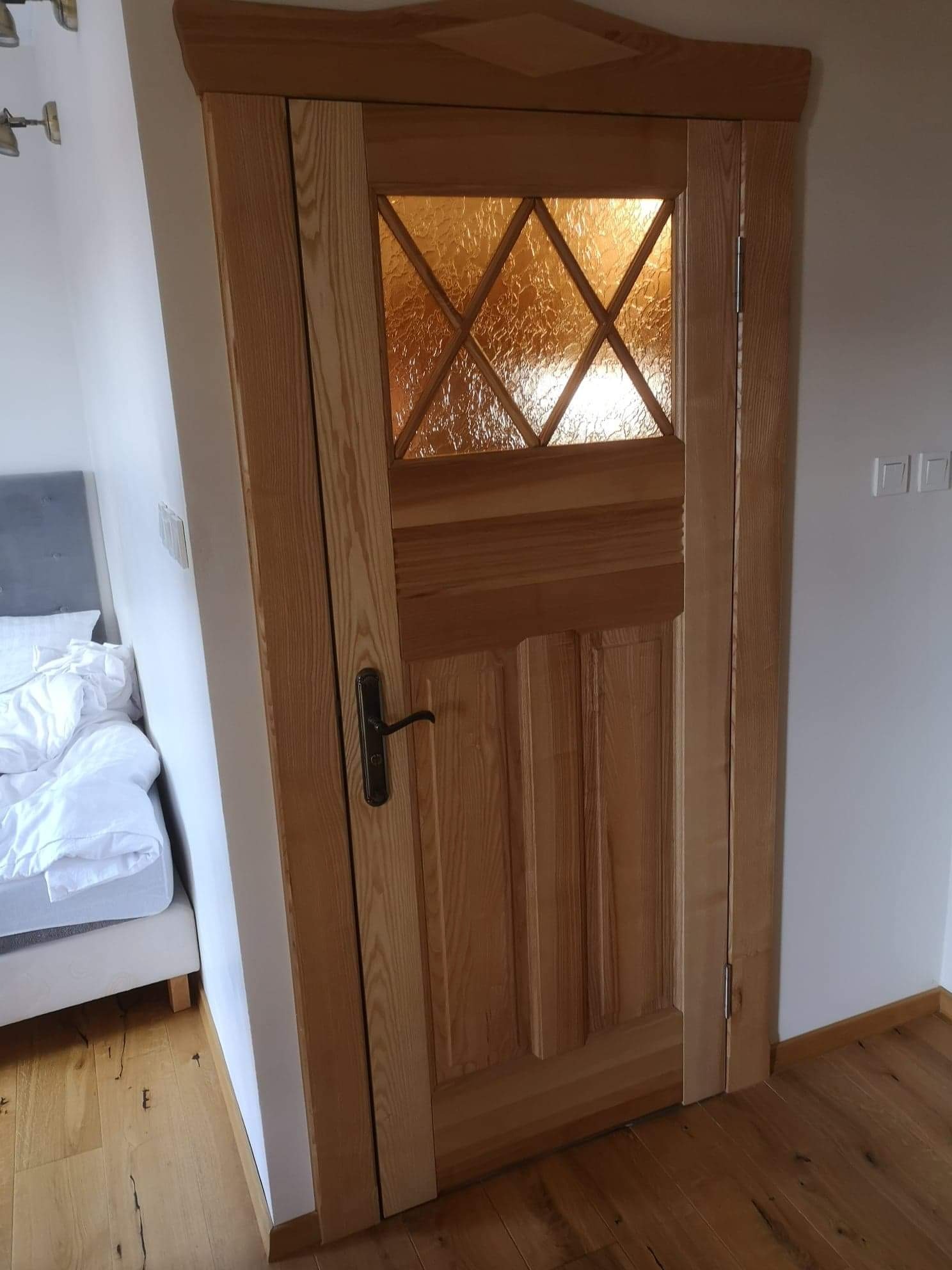 Drewniane drzwi wewnętrzne z szybą o wzorze rombów, widoczne zamek i zawiasy, w tle fragment sypialni z łóżkiem.