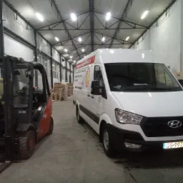 Biały van z logo firmy parkuje obok wózka widłowego w magazynie z rzędem skrzyń w tle.