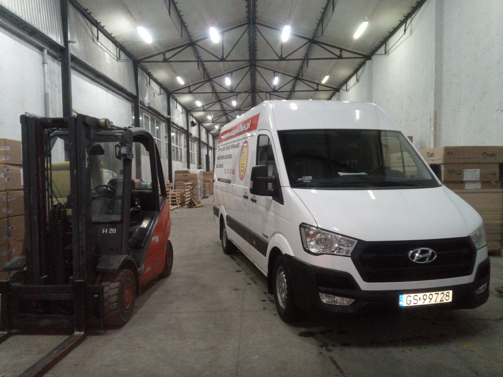 Biały van z logo firmy parkuje obok wózka widłowego w magazynie z rzędem skrzyń w tle.