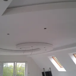 Biały sufit podwieszany z okrągłym elementem dekoracyjnym i otworami na oświetlenie punktowe, widoczne okna dachowe i drabina malarska.