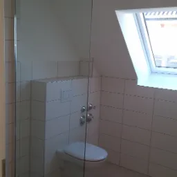 Nowoczesna łazienka na poddaszu z oknem dachowym, białe płytki na ścianach, wisząca toaleta z podtynkową spłuczką i chromowanymi zaworami, kabina prysznicowa ze szklaną ścianką.