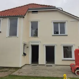 Dwu-kondygnacyjny dom po nałożeniu jasnej elewacji, z nowymi białymi obramowaniami okien i drzwi, widoczna czerwona platforma podnośnikowa i gruz na trawniku.