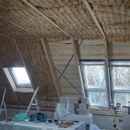Poddasze w trakcie izolacji termicznej, widoczna wełna mineralna między krokwiami, okna dachowe i pracownik w kombinezonie ochronnym.