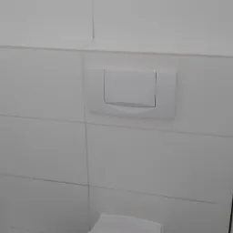 Biała toaleta podwieszana z przyciskiem spłukującym na tle białych, kwadratowych płytek ceramicznych i ciemnoszarej podłogi z płytek.