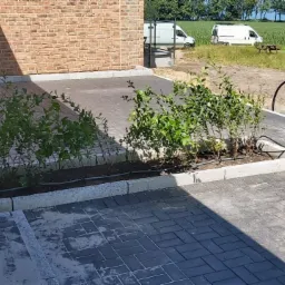 Nowo wybrukowany taras z szarej kostki, z obrzeżami z jasnego kamienia, rabata z młodymi krzewami i systemem nawadniania kropelkowego, na dalszym planie budynek z cegły, dwa białe samochody...