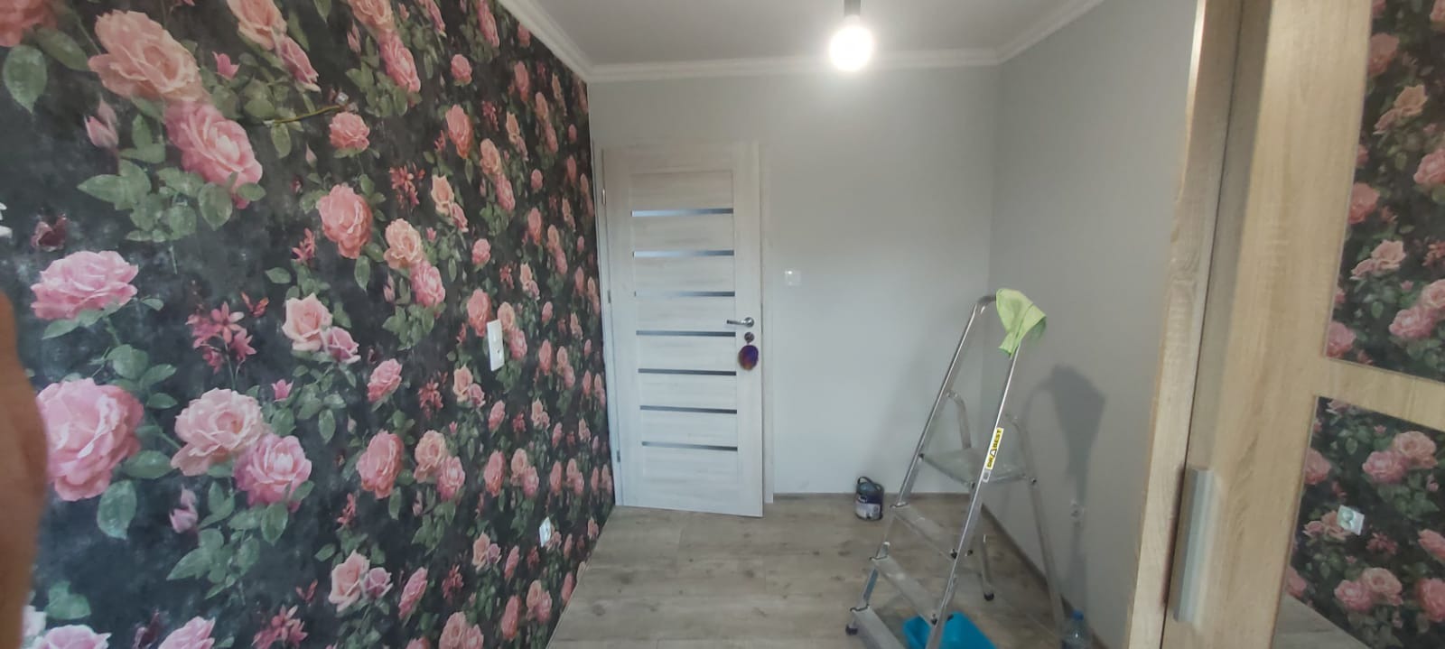 Pokój po tapetowaniu jednej ściany w kwiaty, z drabiną i puszką farby w tle, widoczny fragment szafy z lustrem.