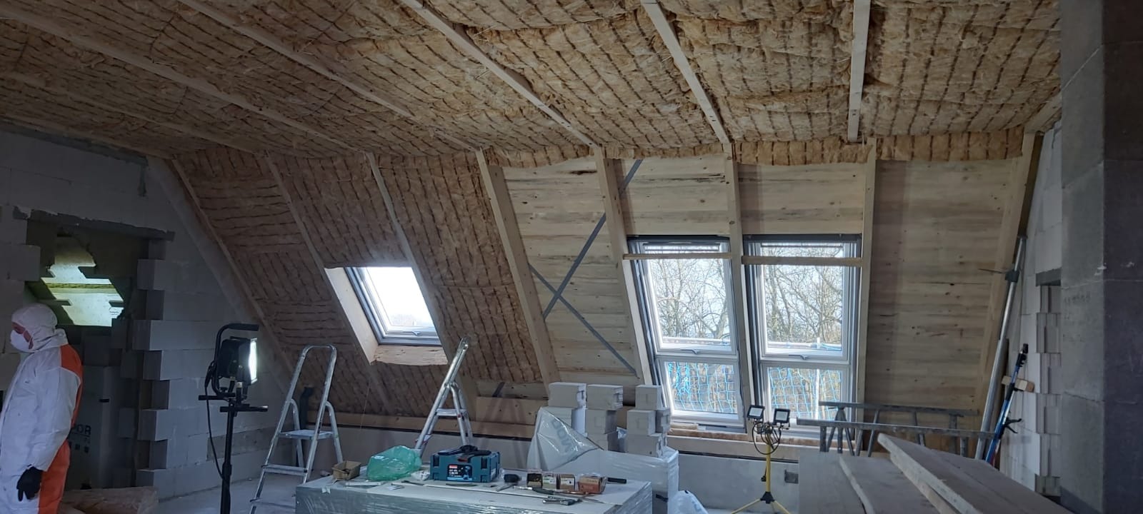 Poddasze w trakcie izolacji termicznej, widoczna wełna mineralna między krokwiami, okna dachowe i pracownik w kombinezonie ochronnym.