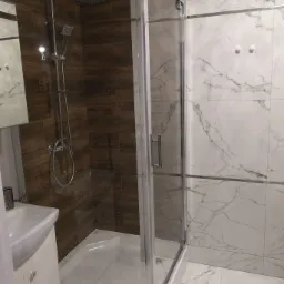 Kabina prysznicowa z chromowaną armaturą na tle płytek imitujących drewno i marmur.