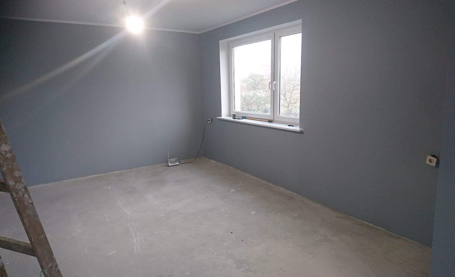 Pomieszczenie po malowaniu ścian na szaro, widoczna drabina malarska, okno z białymi ramami i wystający kabel elektryczny.