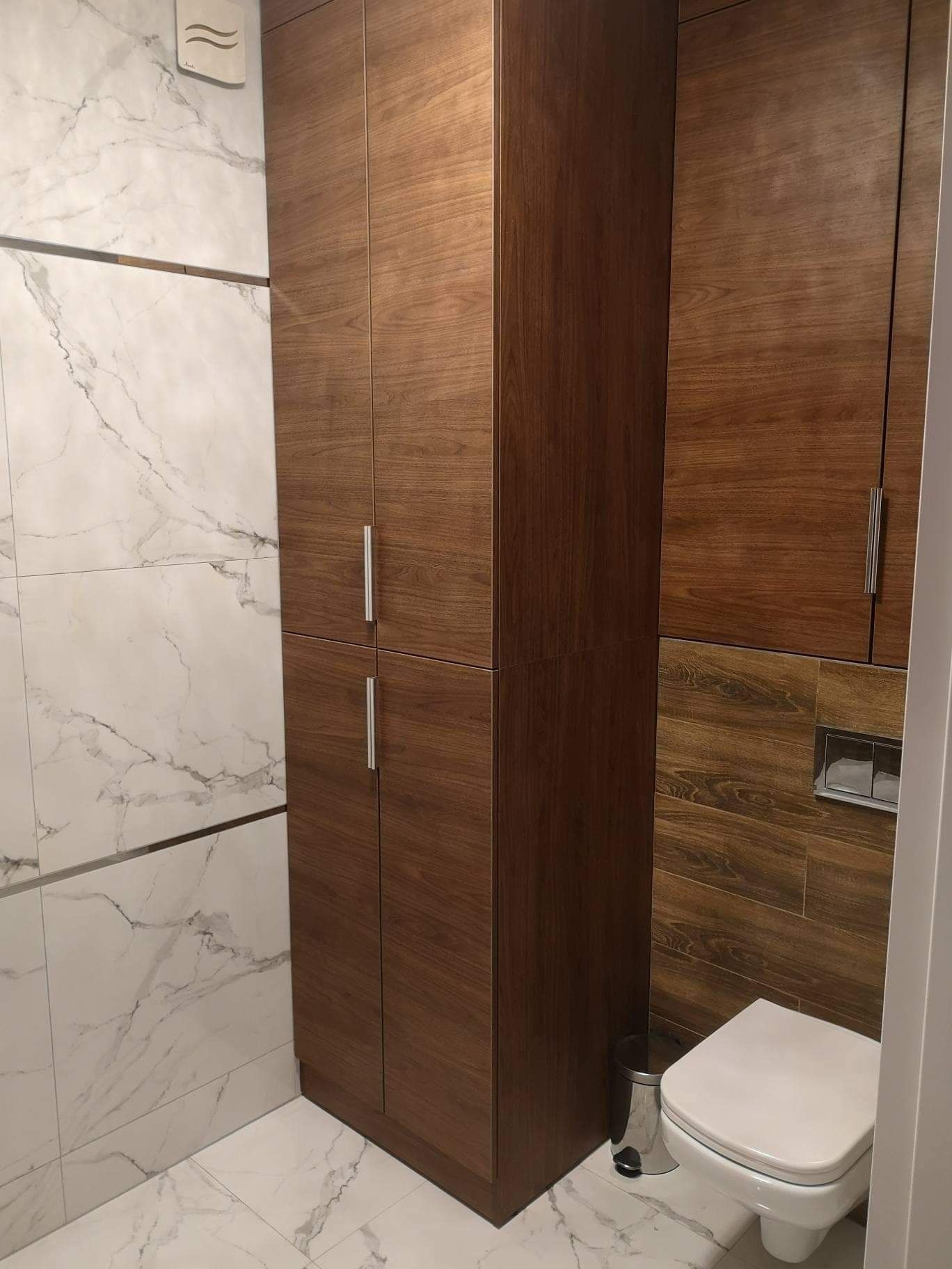 Nowoczesna łazienka z drewnianą zabudową szafkową, marmurowymi płytkami na ścianie i podwieszaną toaletą z ukrytym systemem spłukiwania.