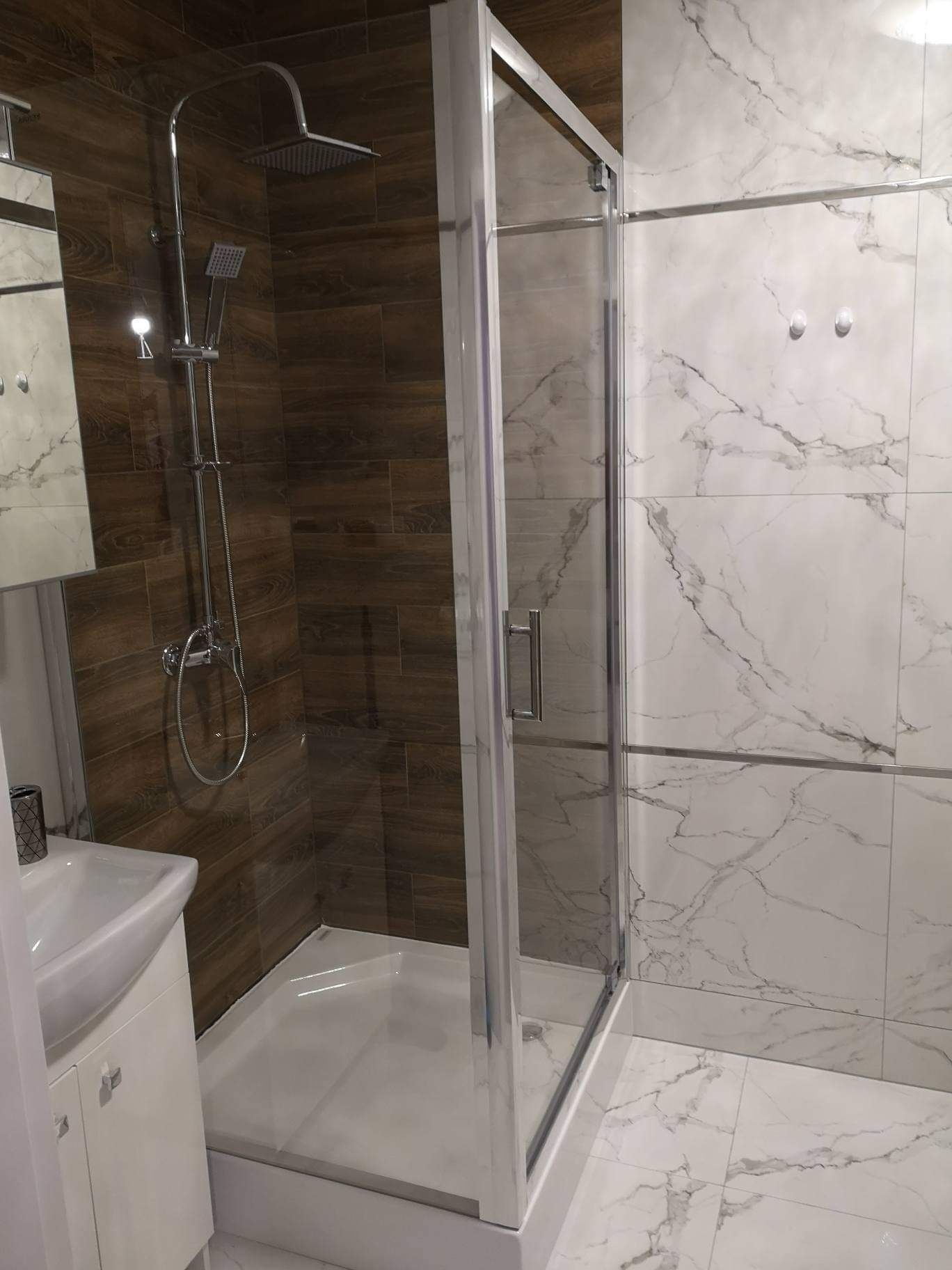 Kabina prysznicowa z chromowaną armaturą na tle płytek imitujących drewno i marmur.