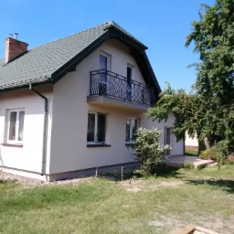 Dwupiętrowy dom z beżową elewacją, zielonym dachem, balkonem z czarną balustradą i ceglanym kominem na tle błękitnego nieba i zieleni drzew.