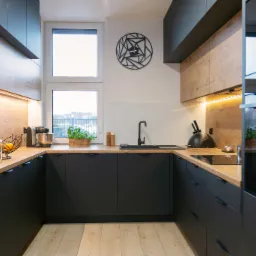 Nowoczesna kuchnia w odcieniach czerni i drewna z minimalistycznym zegarem na ścianie, oświetleniem LED pod szafkami i blatem z owocami i ziołami.