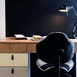 Biurko z drewnianym blatem, kolorowymi szufladami z czarnymi uchwytami i czarną lampką biurkową na tle ciemnej ściany, z widocznym fragmentem krzesła biurowego.