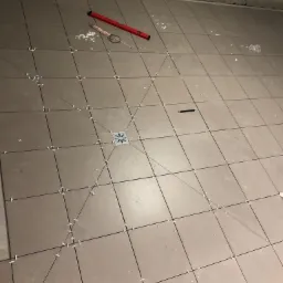 Ułożone szare płytki podłogowe z krzyżykami dystansowymi, widoczny odpływ liniowy, poziomica i ołówek na powierzchni, surowy betonowy sufit w tle.