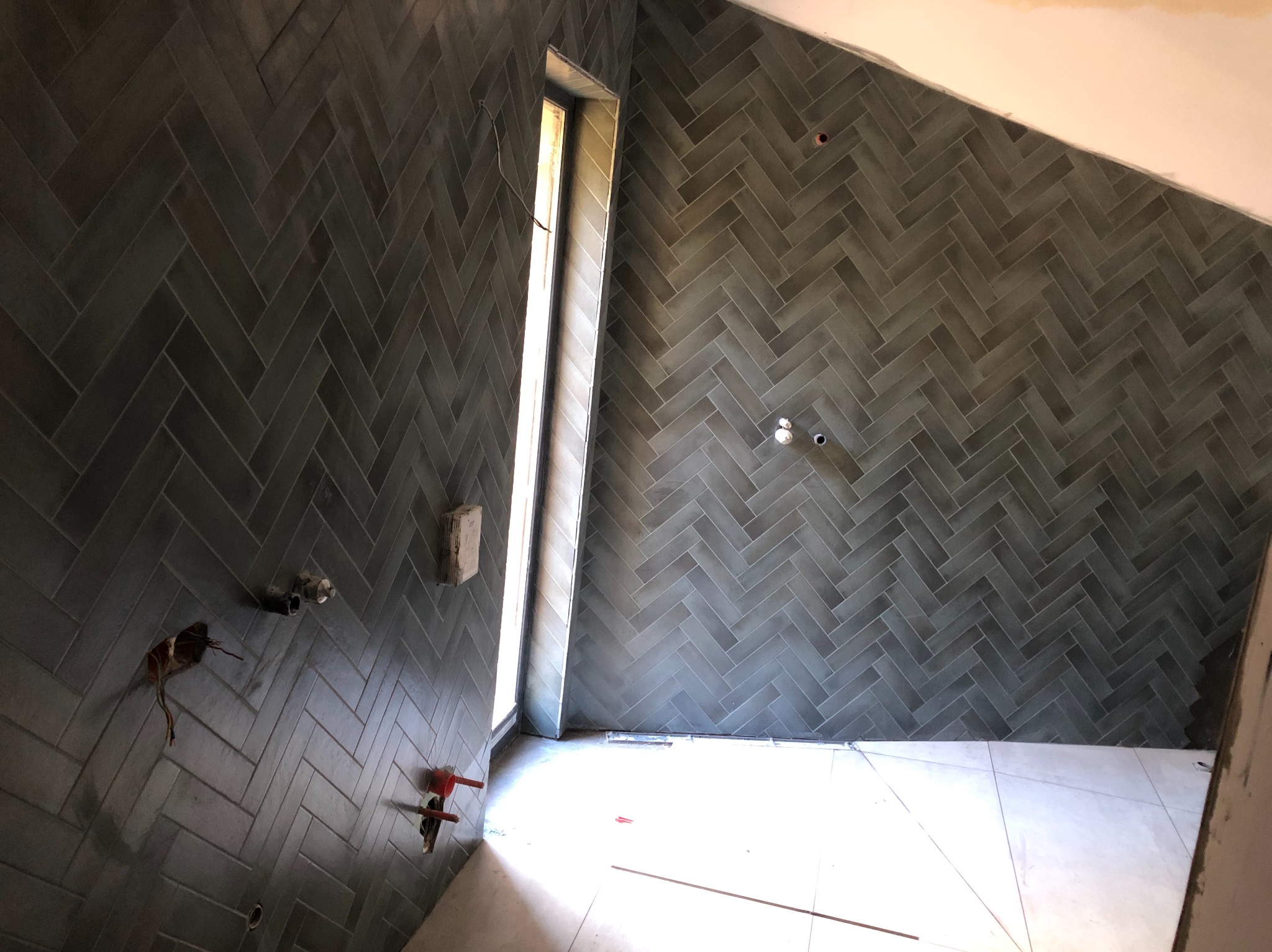 Ściana wyłożona ciemnymi, błyszczącymi płytkami ułożonymi w jodełkę, widoczne niedokończone instalacje i fragment podłogi z jasnych płytek.