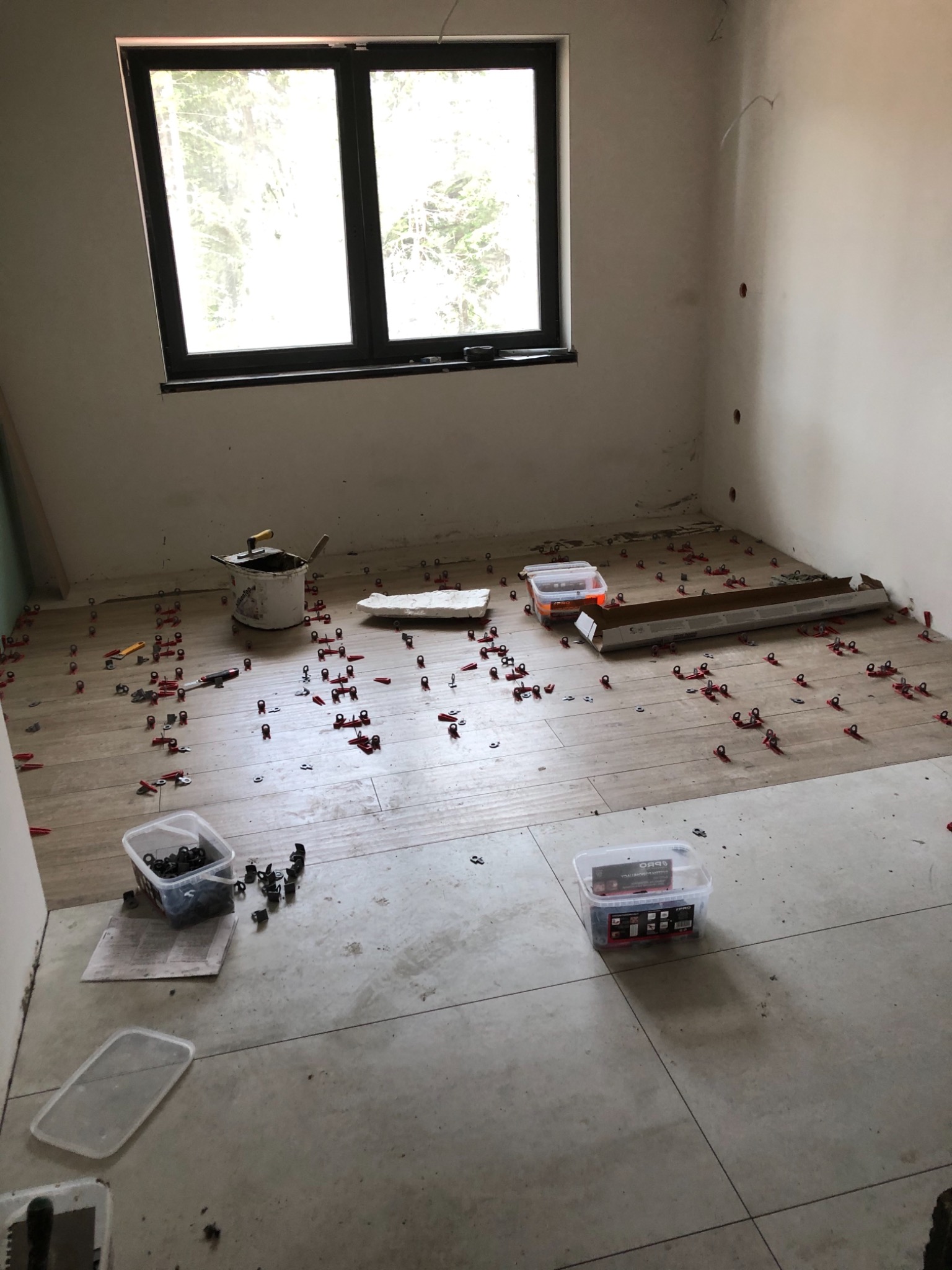 Fragment pokoju w trakcie prac wykończeniowych: częściowo ułożone płytki podłogowe w odcieniach beżu, system poziomowania płytek (klipsy i kliny), wiadro z zaprawą, puszki z akcesoriami, okno...